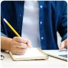 image of a student writing in a spiral notebook with a laptop computer