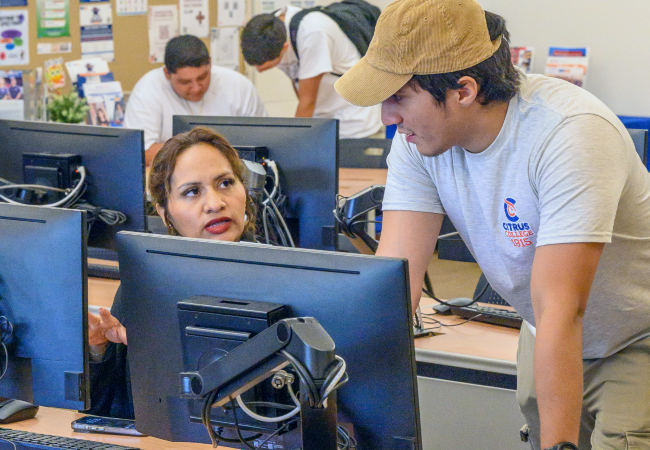 student assistant in a citrus college t-shirt leans over a computer workstation while speaking with a woman seated at another computer in the college's welcome center campus lab