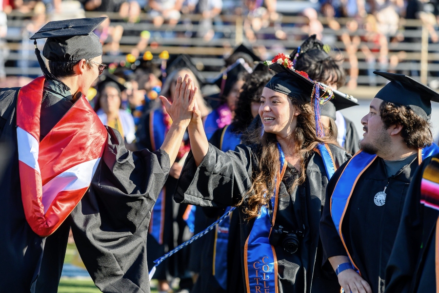 citrus college graduates in caps and gowns celebrate during commencement, with two students exchanging a high five as fellow graduates and guests fill the stadium in the background