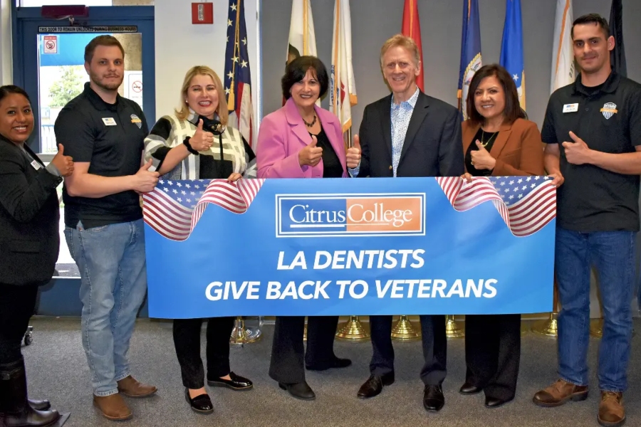 seven people stand together giving thumbs-up while holding a blue citrus college banner reading - la dentists give back to veterans - decorated with american flags; multiple military and government flags are displayed behind the group