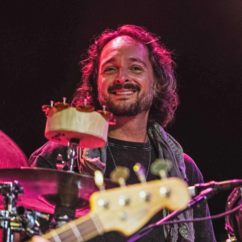 smiling Anthony LoGerfo performs behind a drum kit under colorful stage lighting as a guitarist plays in the foreground