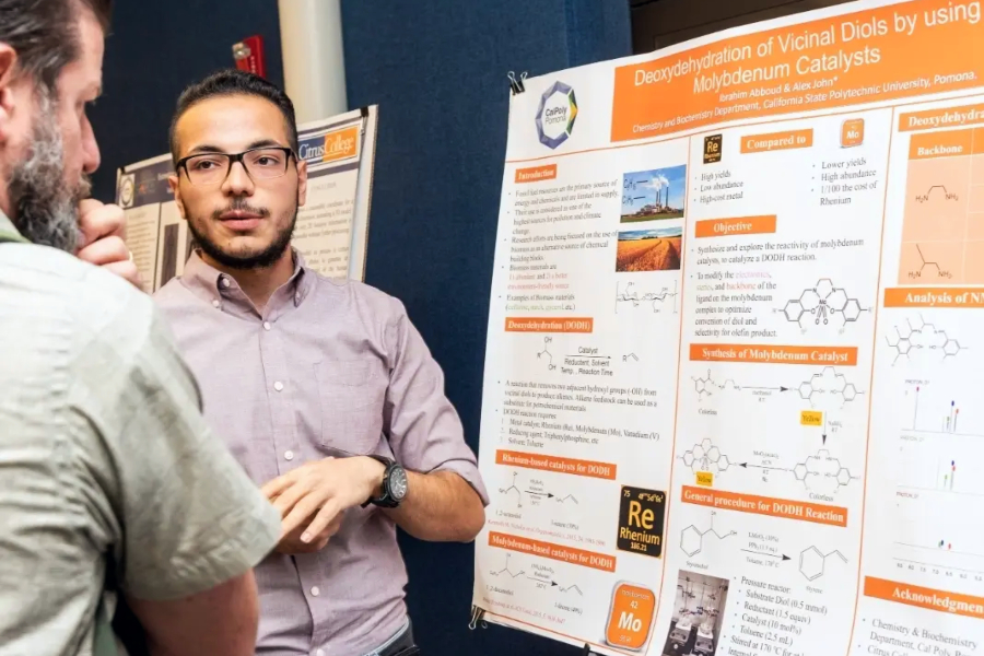 While speaking with an attendee, student Ibrahim Abboud student presents his research beside his scientific poster titled Deoxydehydration of Vicinal Diols by Using Molybdenum Catalysts, featuring chemical structures, diagrams, charts and research findings