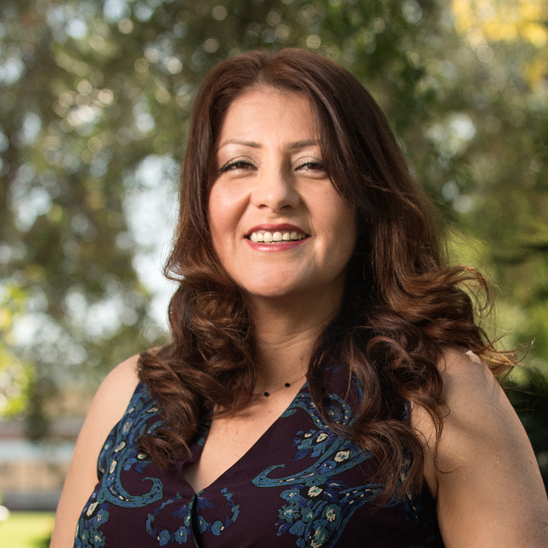 a smiling woman with long, curled brown hair poses outdoors against a softly blurred background of trees